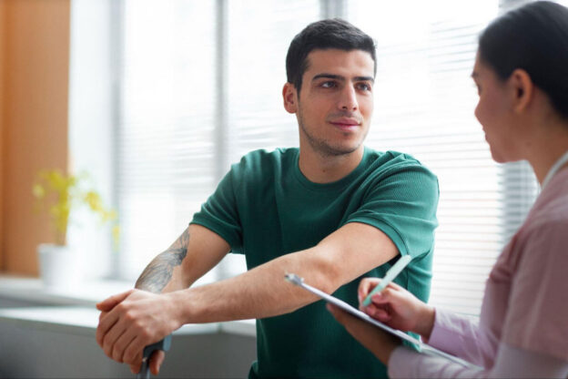 A young tattooed man in a green t-shirt leans forward attentively while a healthcare provider in pink scrubs takes notes on a clipboard during a clinical consultation.