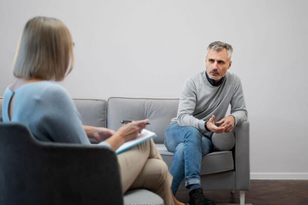 A man sits on a couch with clasped hands speaking thoughtfully to a therapist taking notes during a one-on-one counseling session.