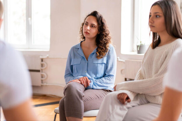 A young woman with curly hair listens attentively while seated in a circle of chairs during a group therapy session.