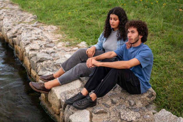 A young man and woman sit thoughtfully on a rocky stone shoreline beside the water, with the woman resting a comforting hand on his shoulder.