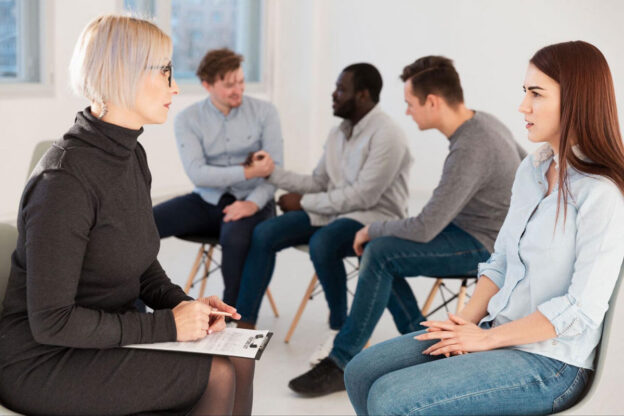 A counselor leads a group therapy session as participants talk in a circle of chairs.
